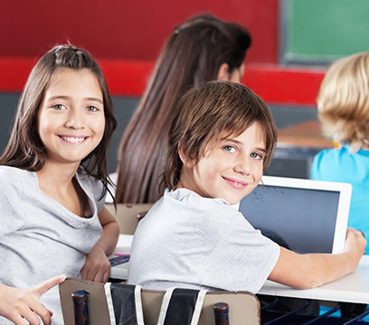 Eine Schülerin und ein Schüler sitzen an einem Tisch mit einem Laptop und drehen sich lächelnd zur Kamera.